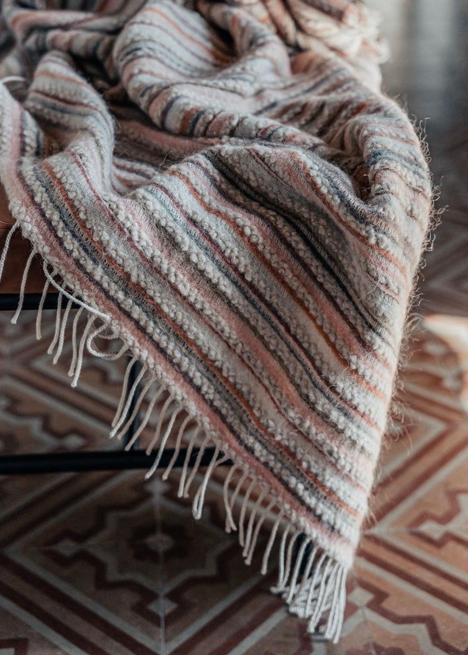 Gestreifte Decke mit feinen Fransen in warmen Naturtönen, drapiert auf einer bank und darunetr historische Fliesen.