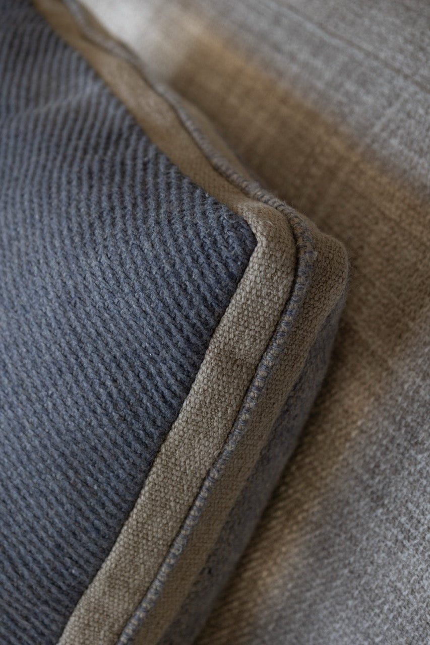 Martin Gabriel Dekokissen Modell Blue wave calm in leinen sichtbar als detailaufnahme schräg von der siet die rückseite im vordergrund in etwas dunklerem blau leinen mit bordüre in beige und mittlekeder in blauem leinen