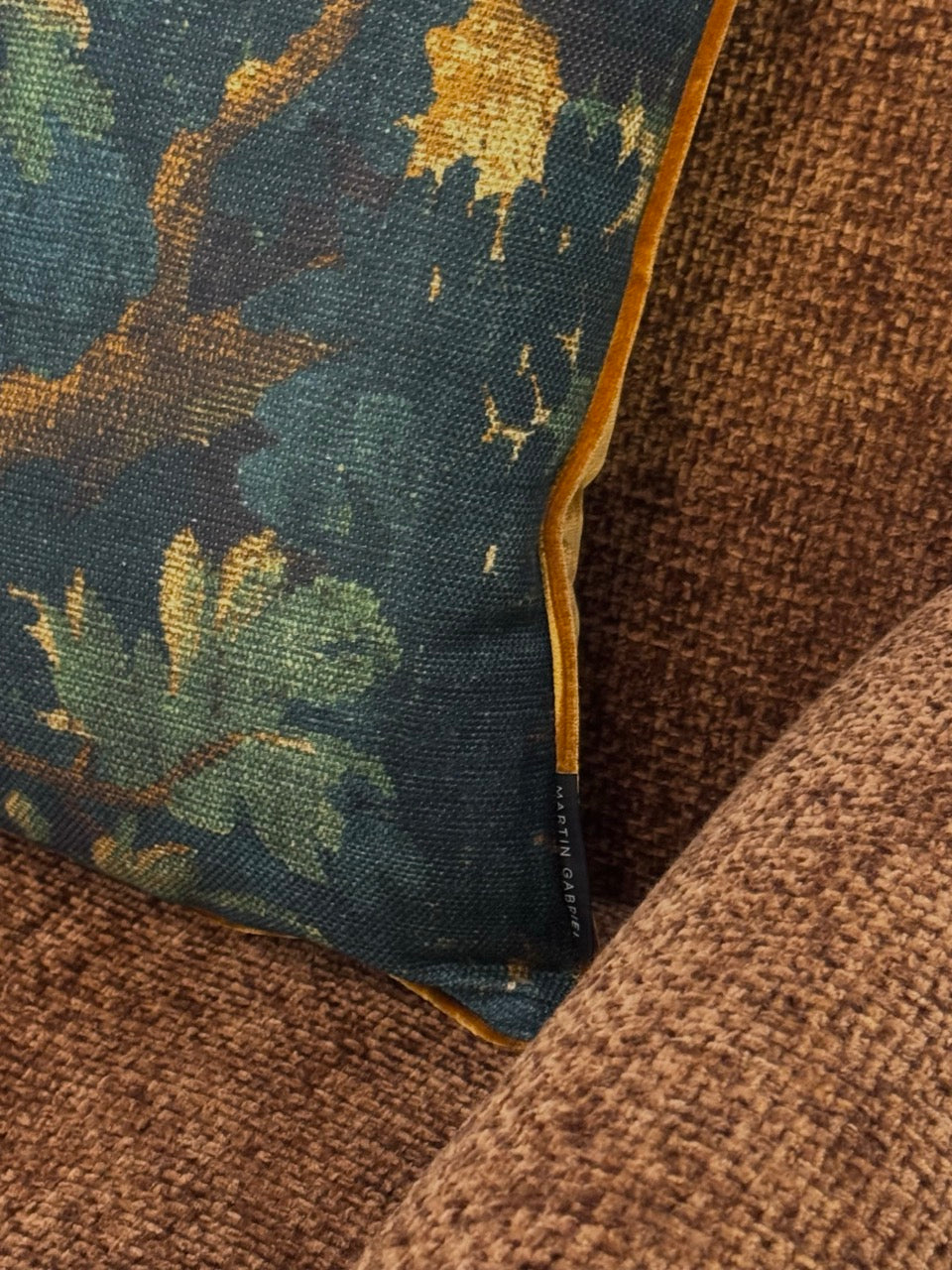 Martin Gabriel Designkissen modell schwarzwald mit goldigen velour keder auf rostbraunem sessel liegend mit label an der seite 