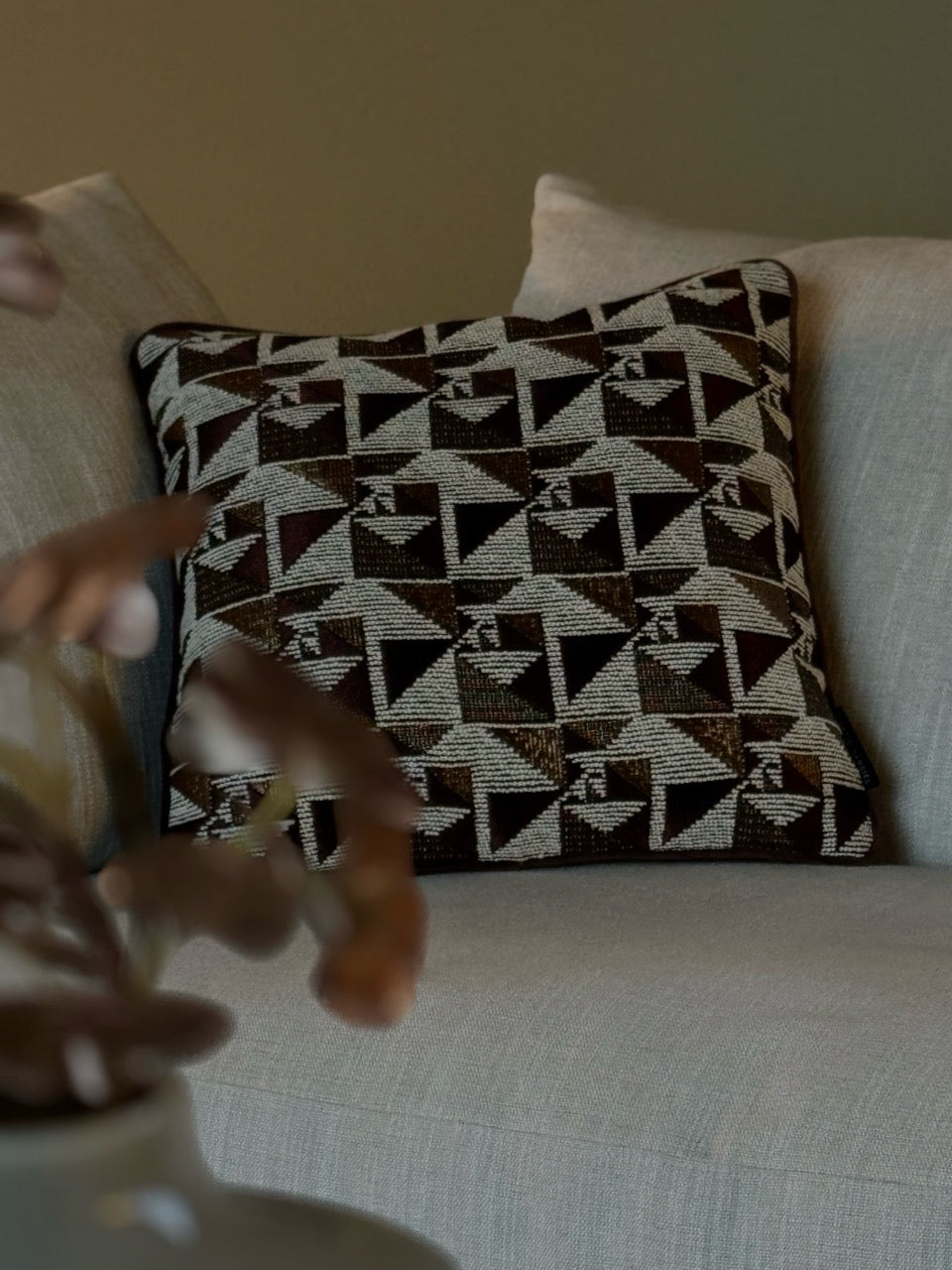 Deko-Kissen in braun-gold-bronze farbenen gometrischen Design auf einem Sofa, mit unscharfer Blume im Vordergrund