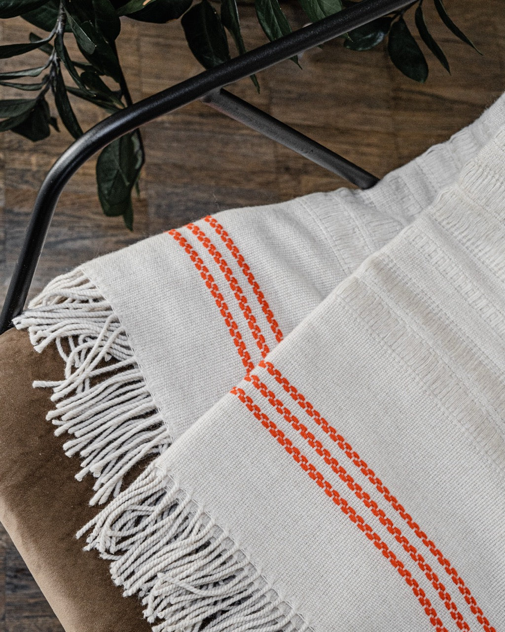 Martin Gabriel Wohndecke Modell Ainu, beige mit fransen und drei dezenten ornagenen streifen drapiert auf einem stuhl