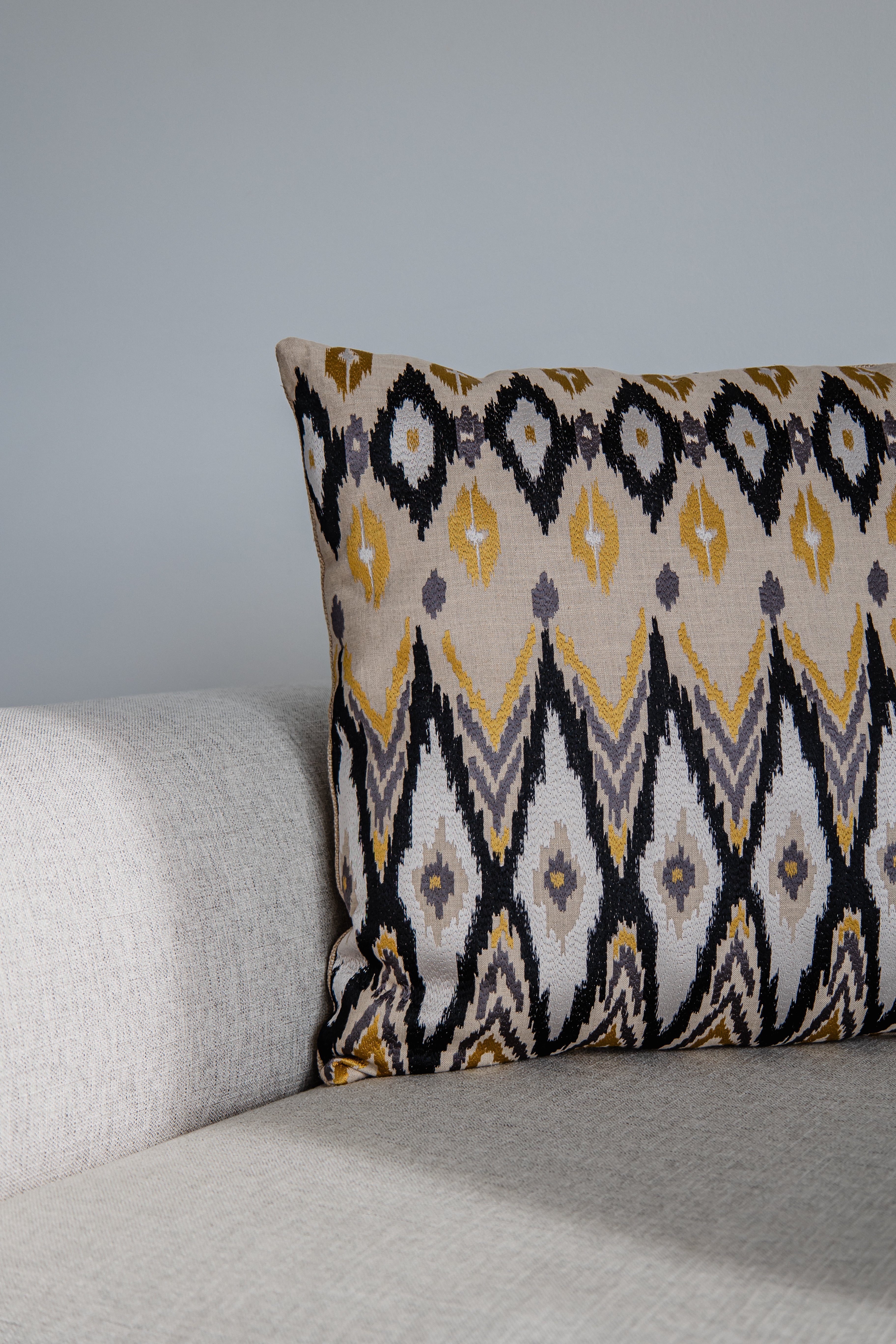 Ikato-Kissen mit geometrischem Muster in Beige, Gelb, Schwarz und Grau, seitlich auf hellem Sofa fotografiert.